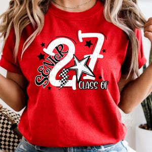 Könnte beinhalten: Ein rotes T-Shirt mit dem Text "SENIOR 27 CLASS OF" in Weiß und Schwarz. Das Design umfasst einen Stern, einen Blitz und kleine schwarze Sterne. Das T-Shirt hat einen Rundhalsausschnitt.
