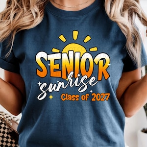 Könnte beinhalten: Marineblaues T-Shirt mit dem Schriftzug "SENIOR sunrise" in Orange und Weiß, mit einem gelben Sonnendesign. Der Text "Class of 2027" befindet sich unter dem Wort "sunrise".