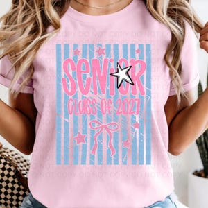 Könnte beinhalten: Ein hellrosa T-Shirt mit dem Aufdruck "SENIOR Class of 2027" in Rosa und Weiß. Das Design umfasst blaue vertikale Streifen, Sterne und eine Schleife. Das T-Shirt hat kurze Ärmel.