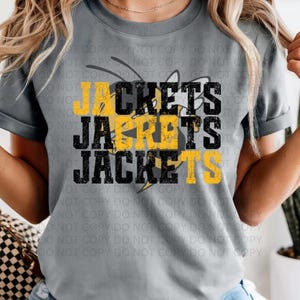 May include: Gray t-shirt with the word "JACKETS" in black and yellow block letters. The word is stacked three times, with a stylized bee graphic incorporated into the design. The shirt has short sleeves.