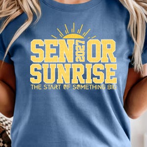 Könnte beinhalten: Blaues T-Shirt mit gelbem Aufdruck "SENIOR SUNRISE 2027" und einem Sonnen-Grafik. Der Satz "THE START OF SOMETHING BIG" steht darunter. Ein auffälliges, farbenfrohes Design für den Abschluss.