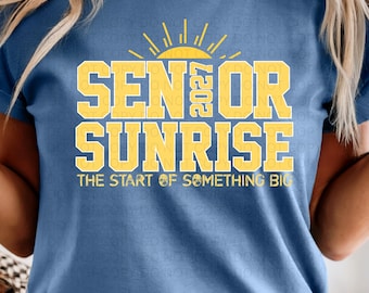 Könnte beinhalten: Blaues T-Shirt mit gelbem Aufdruck "SENIOR SUNRISE 2027" und einem Sonnen-Grafik. Der Satz "THE START OF SOMETHING BIG" steht darunter. Ein auffälliges, farbenfrohes Design für den Abschluss.