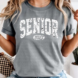 K&ouml;nnte beinhalten: Ein graues T-Shirt mit dem Wort "SENIOR" in gro&szlig;en, wei&szlig;en, abgenutzten Buchstaben. Unter dem Wort "SENIOR" steht das Jahr "2022" in einem wei&szlig;en Oval. Das Shirt ist ein l&auml;ssiger Stil.