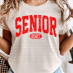 K&ouml;nnte beinhalten: Cremefarbenes T-Shirt mit dem Wort "SENIOR" in verblichenen roten Buchstaben. Unter dem Wort "SENIOR" steht die Zahl "2027" in einem roten Oval. Das T-Shirt ist ein l&auml;ssiger Stil.