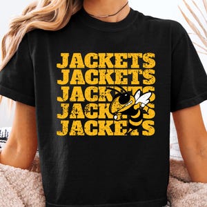 May include: A black short-sleeved t-shirt featuring the word JACKETS printed in a distressed yellow font across five horizontal lines. A graphic of a yellow and black hornet with white wings is positioned over the text on the right side.