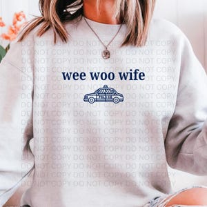 May include: A light gray sweatshirt with the text "wee woo wife" in navy blue, along with a navy blue police car graphic. The sweatshirt is a casual style.