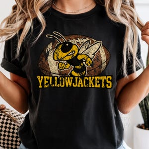 May include: Black t-shirt featuring a yellow and black cartoon bee with a football in the background. The text "YELLOWJACKETS" is printed in yellow below the bee and football graphic. The shirt is a comfortable, casual style.