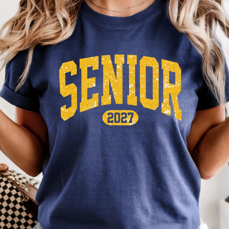 K&ouml;nnte beinhalten: Marineblaues T-Shirt mit dem Wort "SENIOR" in vergilbten gelben Blockbuchstaben. Unter dem Wort "SENIOR" steht die Zahl "2027" in einem gelben Oval. Das T-Shirt hat kurze &Auml;rmel.