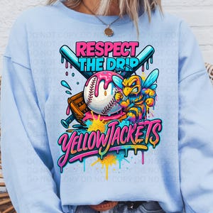 May include: Light blue sweatshirt with a colorful graphic. The graphic features a baseball with pink frosting, crossed bats, and a cartoon bee. Text reads "Respect the Drip" and "Yellowjackets".