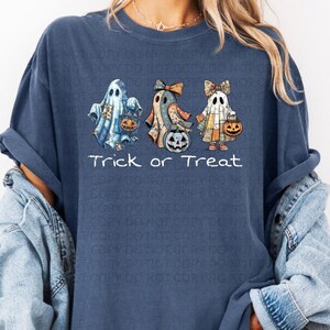 May include: A dark blue t-shirt featuring a graphic of three ghosts wearing patterned fabric sheets and bows, each holding a jack-o'-lantern bucket. Below the ghosts, the text Trick or Treat is printed in a white, simple sans-serif font.