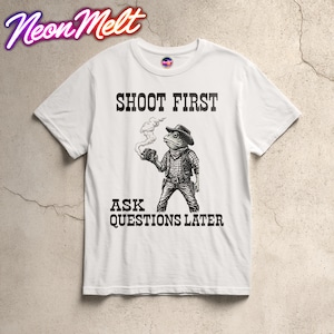 May include: White t-shirt with a graphic of a frog wearing a cowboy hat and vest, holding a smoking gun. The text "SHOOT FIRST" and "ASK QUESTIONS LATER" are printed above and below the frog illustration.