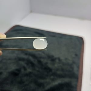 Op de afbeelding: Een doorschijnende, ovale edelsteen van melkachtige witte kleur wordt vastgehouden door een pincet. De edelsteen heeft een glad, gepolijst oppervlak. De pincet is zilverkleurig.