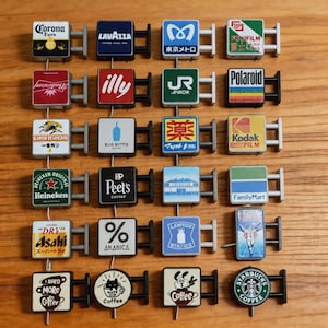 Könnte beinhalten: Eine Sammlung von Miniatur-Schildrepliken mit Logos verschiedener Marken, darunter Corona, Lavazza und Starbucks. Jedes Schild hat eine rechteckige oder kreisförmige Form mit einem Metallrahmen. Die Schilder sind auf einer Holzoberfläche angeordnet.