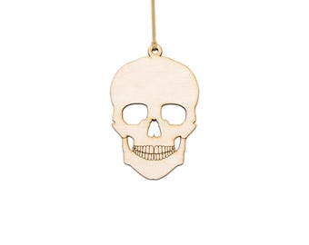Pode incluir: Um ornamento de caveira de madeira com cavidades oculares, cavidade nasal e uma fileira de dentes. Suspenso por um fio fino castanho claro. O ornamento é feito de madeira clara.