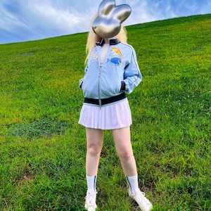 Puede incluir: Una chaqueta deportiva azul claro con cremallera y un gráfico de un pez cirujano y un arcoíris en el pecho, combinada con una minifalda plisada blanca, calcetines blancos con rayas azules y zapatillas blancas. La chaqueta tiene detalles negros.