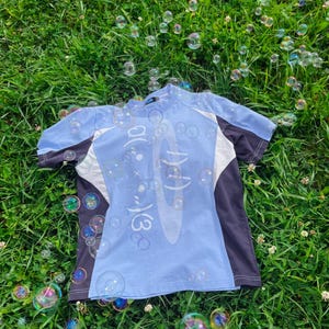 Op de afbeelding: Een lichtblauw t-shirt met korte mouwen, donkergrijze zijpanelen, witte driehoekige accenten en een centrale grafische print met witte tekst en symbolen. Het shirt heeft een ronde hals en zichtbare witte stiksels langs de zijnaden.