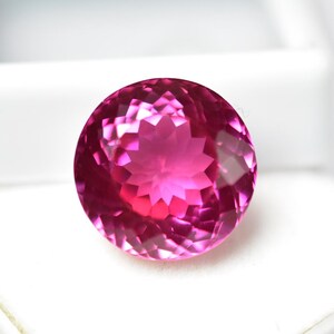 May include: A faceted, round gemstone with a vibrant pink hue. The gem's intricate cuts create a starburst pattern in the center, reflecting light. The gemstone is set against a white background, highlighting its color and clarity. This is a gemstone.