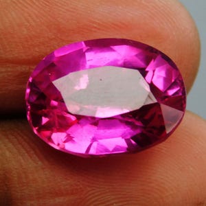 Puede incluir: Una gema rosa intenso facetada, de forma ovalada. La gema tiene un corte brillante con múltiples facetas que reflejan la luz. La piedra está sujeta en la mano de una persona.