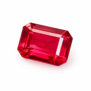Può includere: Una vibrante gemma di rubino a taglio rettangolare. La gemma è di un colore rosso intenso e saturo con sfaccettature che riflettono la luce. La forma è a taglio smeraldo, con un corpo rettangolare e angoli smussati, su sfondo bianco.