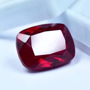 May include: A faceted, cushion-cut ruby gemstone. The gem is a deep, saturated red colour with multiple reflective facets. The shape is a rounded rectangle, showcasing the stone's brilliance.