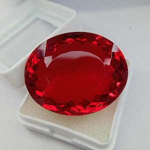 May include: A large, oval-shaped, faceted gemstone with a vibrant, deep red colour. The stone features a complex pattern of triangular and diamond-shaped facets that reflect light across its surface. It is displayed resting inside a white plastic box.
