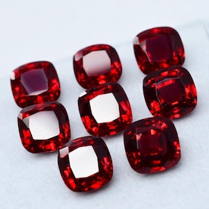 May include: Eight cushion-cut, deep red gemstones are arranged on a white surface. Each gem has a square shape with rounded corners and a bright, reflective surface. The gems are a rich, saturated red color.