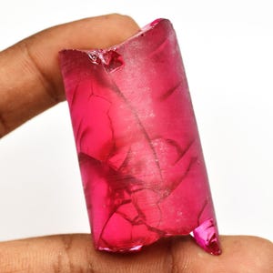 May include: A vibrant, translucent, cylindrical gemstone in a deep magenta hue. The stone has visible internal fractures and a slightly irregular shape. The gem is held against a white background, highlighting its color and clarity.