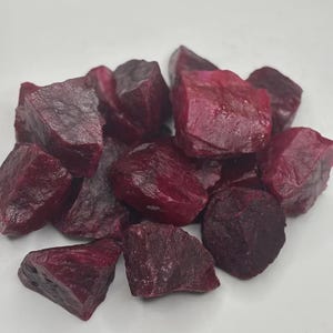 May include: A collection of raw, unpolished ruby gemstones with a deep, saturated magenta-red color. Each stone features irregular, jagged edges and a natural, textured surface with varying facets that catch the light, showcasing a rich, opaque appearance.