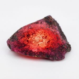May include: A rough-cut, natural ruby gemstone with a vibrant red and pink hue. The stone has a textured surface with dark, almost black, areas contrasting with the bright, glowing center. The overall shape is irregular, showcasing its raw, unpolished state.