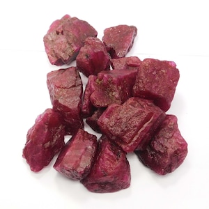 Puede incluir: Una colección de piedras preciosas de rubí en bruto y sin pulir con un color rojo magenta profundo y saturado. Cada piedra presenta una textura rugosa e irregular con facetas naturales, bordes afilados y estructuras cristalinas variadas.