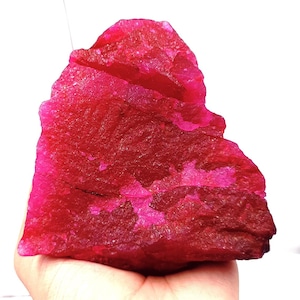 May include: A rough, uncut ruby gemstone with a deep red hue and areas of vibrant pink. The natural stone has a textured surface and irregular shape, showcasing its raw, unrefined beauty. The gemstone is held in a hand.