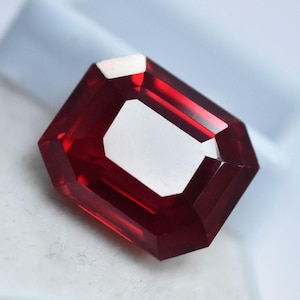 May include: A rectangular-cut, deep red gemstone with a bright, white interior. The gem has a polished surface and is set against a soft, white background. The gemstone's facets reflect light, creating a brilliant appearance.