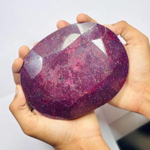 Puede incluir: Una gran gema de rubí facetada, de forma ovalada y color rojo-púrpura intenso. La piedra se sostiene con dos manos sobre un fondo blanco. La superficie de la gema refleja la luz, mostrando sus múltiples facetas y patrones internos. Ideal para joyería.