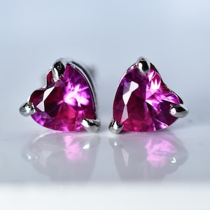 Può includere: Un paio di orecchini a lobo a forma di cuore. Ogni orecchino presenta una gemma rosa vibrante sfaccettata, incastonata in una montatura in metallo argentato. Gli orecchini sono esposti su una superficie bianca riflettente.