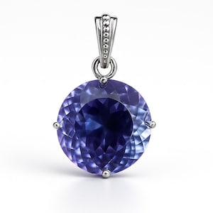 Peut inclure: Pendentif en argent avec une pierre précieuse violette ronde et facettée. La pierre a un centre bleu foncé qui s'estompe en un violet plus clair vers les bords. La bélière en argent est ornée de petits accents ronds.