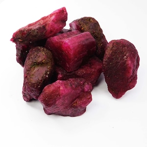 May include: A cluster of raw, unpolished ruby gemstones featuring deep magenta and dark red hues. The stones exhibit irregular, jagged shapes with natural crystalline textures, varying surface facets, and earthy brown mineral inclusions.