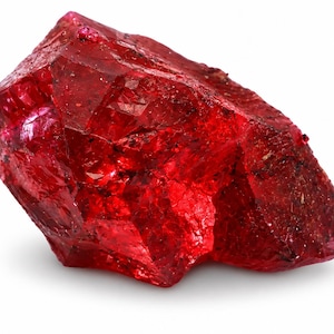 May include: A raw, unpolished ruby gemstone with a deep, vibrant red colour. The stone features a jagged, crystalline structure with multiple natural facets, sharp edges, and small dark inclusions embedded within its translucent, glowing surface.