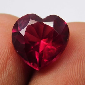 May include: A faceted, heart-shaped gemstone with a deep red hue. The gem's surface reflects light, showcasing its multiple facets and internal structure. The gemstone is a vibrant, eye-catching piece.