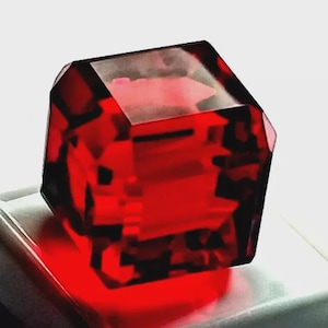 Op de afbeelding: Een doorschijnend, dieprood, veelzijdig kubusobject met scherpe randen en geometrische oppervlakken. Het materiaal lijkt op heldere, gepolijste hars of glas, waardoor licht doorlaat en interne schaduwen en reflecties worden gecreëerd.