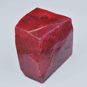 Puede incluir: Una gema de rubí en bruto, facetada, de color rojo intenso. La gema tiene forma rectangular con múltiples facetas, mostrando su textura natural y sin pulir. El color varía ligeramente en toda la piedra.