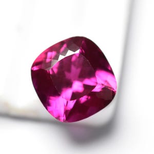 May include: A faceted, cushion-cut gemstone in a vibrant magenta hue. The gem's multiple facets reflect light, creating a sparkling effect. The gemstone is set against a white background, highlighting its color and clarity. This gem is suitable for jewelry.