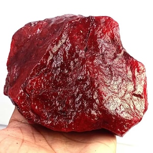Puede incluir: Una gema grande, en bruto, de color rojo intenso con una superficie rugosa y texturizada y bordes irregulares y dentados. La piedra muestra un color carmesí vibrante y saturado, con grietas naturales y facetas reflectantes.