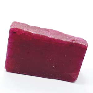 Puede incluir: Una gema en bruto de forma rectangular, de color magenta intenso. La piedra tiene una superficie superior rugosa y texturizada y lados lisos y pulidos. Probablemente sea un espécimen mineral.