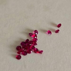 May include: A scattering of small, oval-cut rubies. The gemstones are a rich, deep red hue and are arranged on a pale surface. The rubies, of varying sizes, catch the light, displaying their facets.