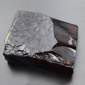 May include: A dark amber-colored, irregularly shaped piece of amber. The amber has a translucent quality with a textured surface on one side and a smooth, glossy surface on the other. The edges are rough and uneven, with some areas appearing chipped.