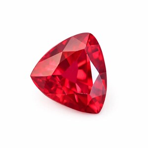 Op de afbeelding: Een gefacetteerde, driehoekige, rode edelsteen. De edelsteen heeft vele facetten die licht reflecteren, waardoor een briljante uitstraling ontstaat. De edelsteen staat tegen een witte achtergrond.