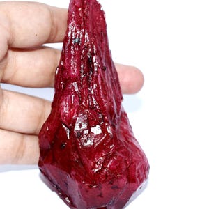 May include: A raw, deep red crystal specimen with a pointed, elongated shape and a textured, multifaceted surface. The stone displays a glossy, translucent appearance with dark inclusions scattered throughout its irregular, jagged crystalline structure.