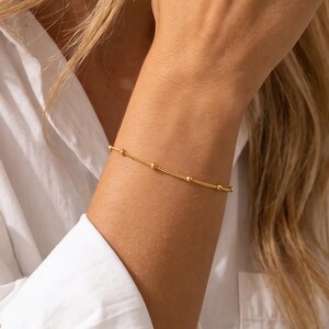 May include: A delicate gold-toned chain bracelet featuring small, evenly spaced spherical beads along the length of the chain. The bracelet is worn on the wrist, showcasing a minimalist design with a thin, polished metal link construction.