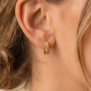 May include: A small gold hoop earring with a rectangular shape and rounded edges. The earring features a polished metallic finish and is worn in the earlobe, showcasing a simple, clean design that sits flush against the skin of the ear.