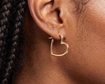 May include: A gold-toned hoop earring shaped like an open heart outline. The earring features a smooth, polished metallic finish and is secured through the earlobe with a thin wire post, creating a simple and clean silhouette for the jewelry piece.